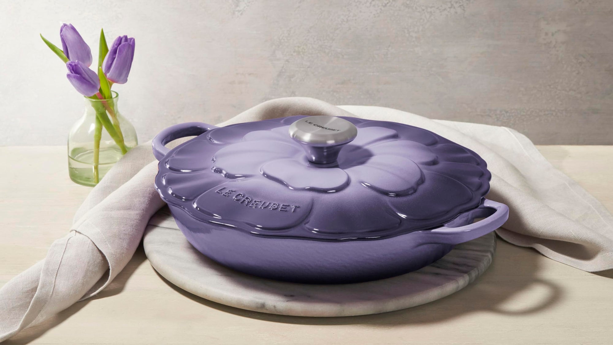 Purple cast iron cookware with floral design on lid on a marble surface with a white napkin and purple tulips.