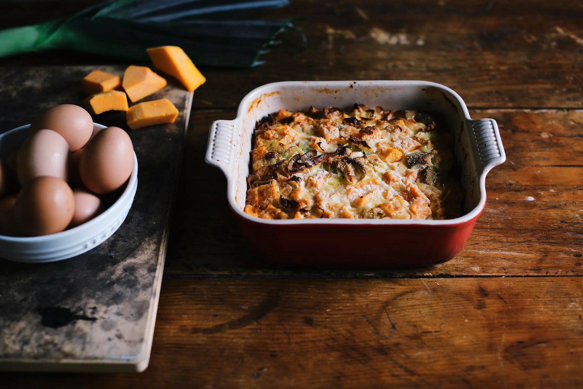 Cerise Heritage 3 Qt Square Dish with a casserole inside displayed on a wooden surface next to eggs and cheese