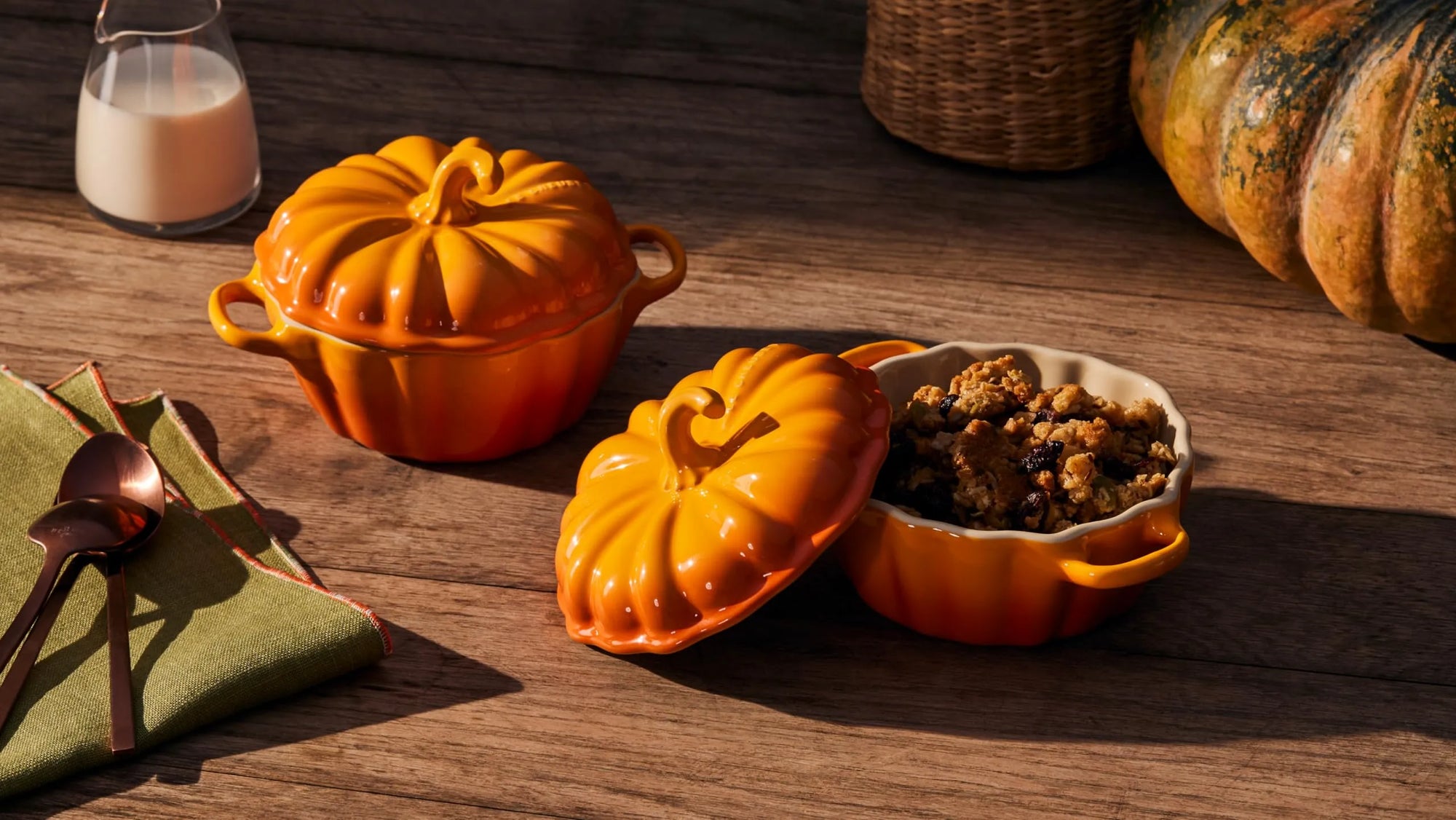 Two orange pumpkin-shaped ceramic pots on a rustic wooden table, one closed and one open with its lid off, showing cooked food inside. A folded green napkin with spoons, a glass of milk, and part of a wicker basket are also visible in the scene.