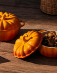 Two orange pumpkin-shaped ceramic pots on a rustic wooden table, one closed and one open with its lid off, showing cooked food inside. A folded green napkin with spoons, a glass of milk, and part of a wicker basket are also visible in the scene.