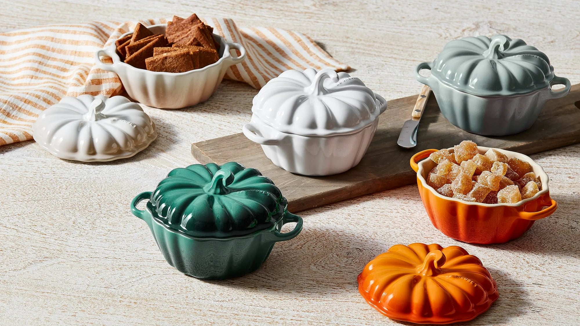 A set of pumpkin-shaped ceramic casserole dishes in white, teal, orange, and gray, shown with their lids off and filled with food on a light wood surface.