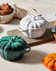A set of pumpkin-shaped ceramic casserole dishes in white, teal, orange, and gray, shown with their lids off and filled with food on a light wood surface.