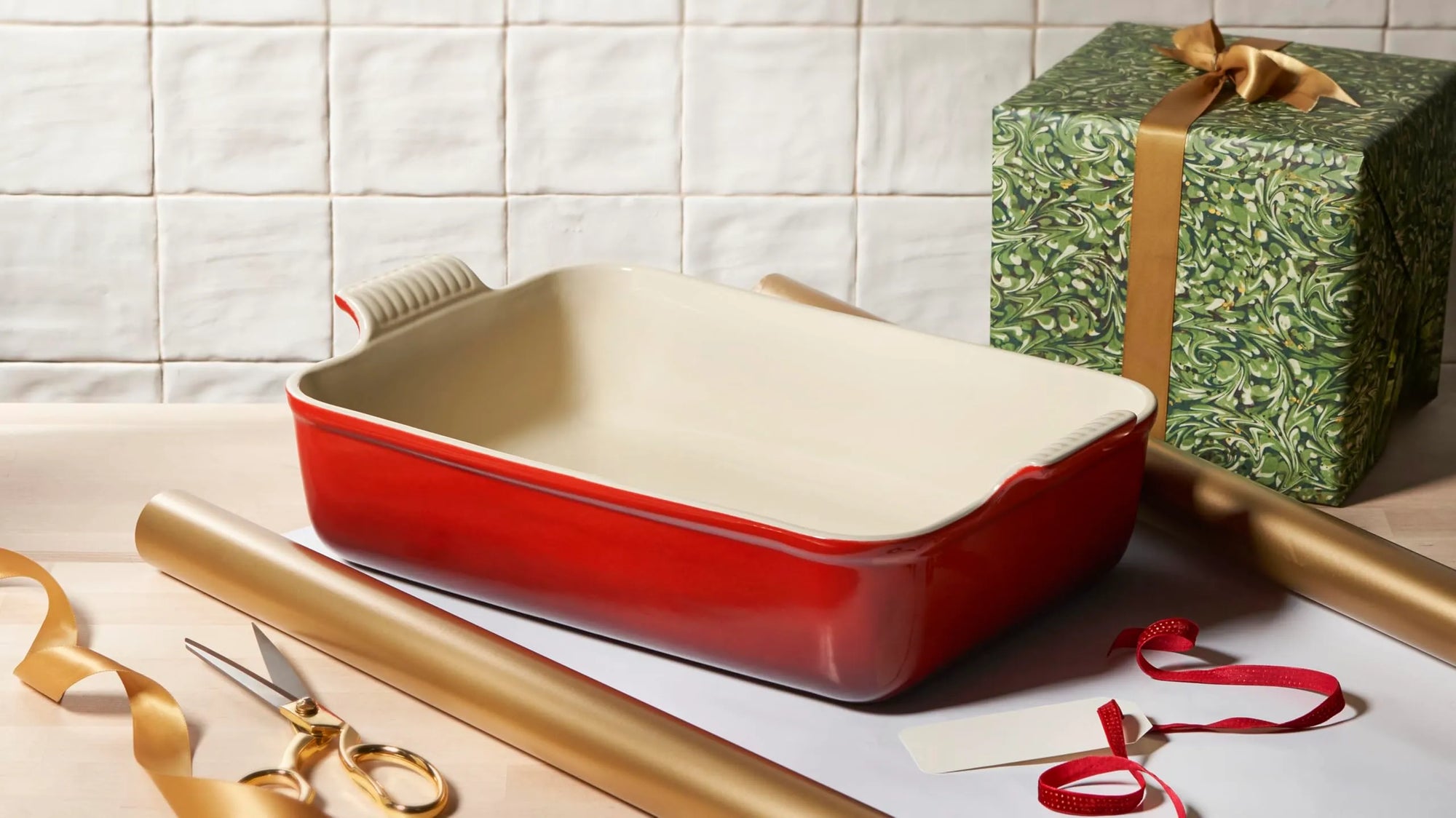 A red rectangular baking pan with a cream-colored interior sits on a tabletop surrounded by gift-wrap materials, including gold ribbon, gold tape, scissors, and a green patterned gift box with a gold ribbon bow.