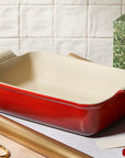 A red rectangular baking pan with a cream-colored interior sits on a tabletop surrounded by gift-wrap materials, including gold ribbon, gold tape, scissors, and a green patterned gift box with a gold ribbon bow.