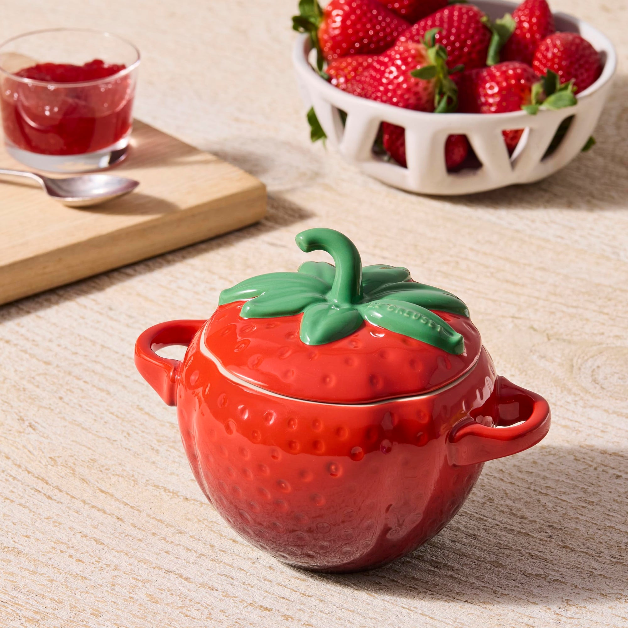 Red strawberry-shaped container with green lid on a wooden surface, next to a bowl of strawberries and a glass of red liquid.