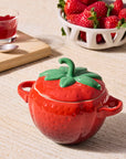 Red strawberry-shaped container with green lid on a wooden surface, next to a bowl of strawberries and a glass of red liquid.