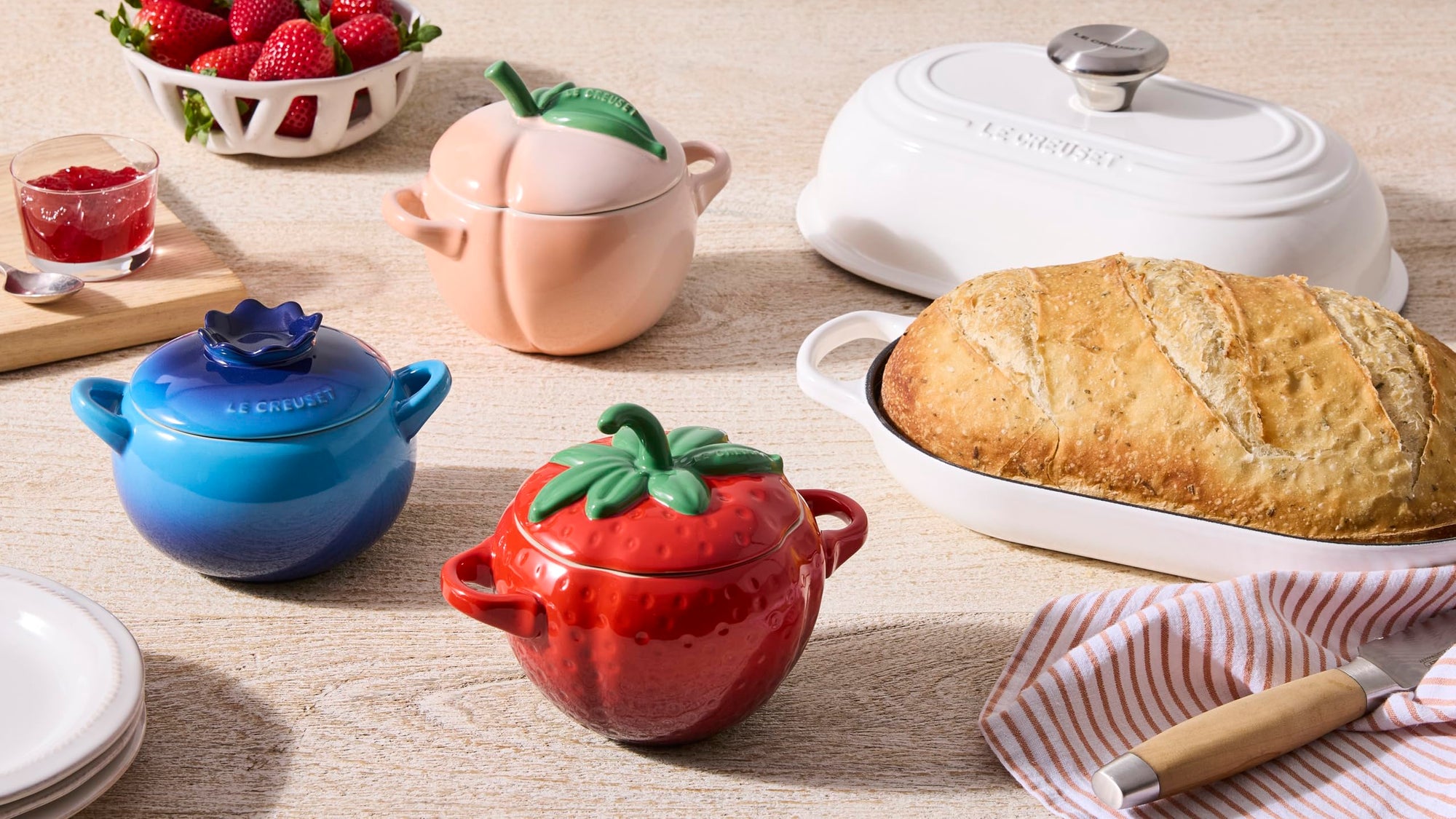 Colorful ceramic pots shaped like strawberries, peaches, and blueberries on a wooden table with bread and berries.