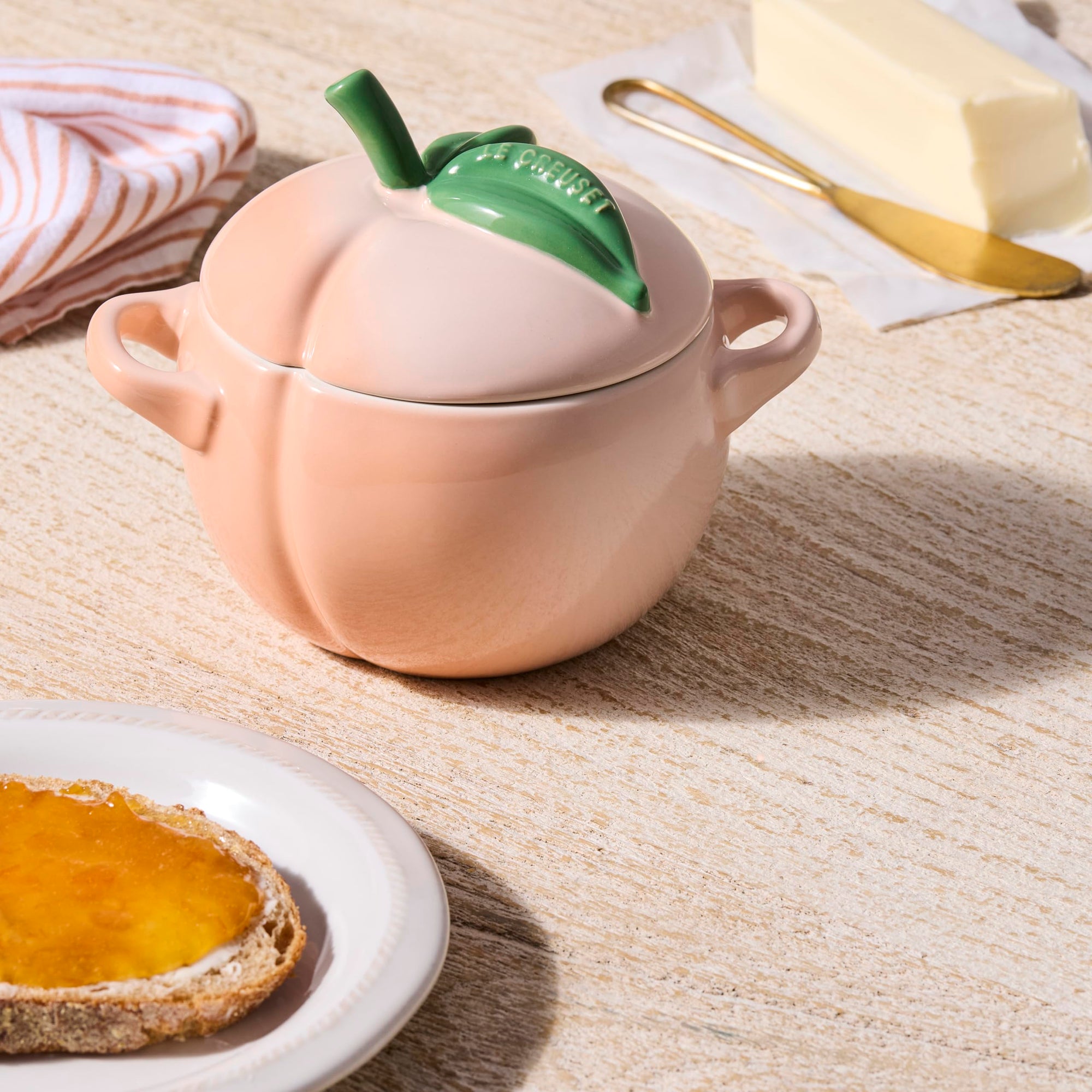 Peach-shaped ceramic container with a green leaf next to bread, butter, and butter knife on a wooden surface