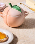 Peach-shaped ceramic container with a green leaf next to bread, butter, and butter knife on a wooden surface