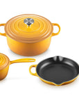 A three-piece cookware set in a bright yellow-orange enamel finish, including a lidded pot, a smaller lidded pot, and a skillet with a black interior, shown on a white background.
