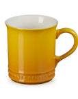 A Le Creuset ceramic mug in a vibrant mustard yellow color, featuring a glossy finish, a curved handle, and the embossed Le Creuset brand name at the base, set against a plain white background.