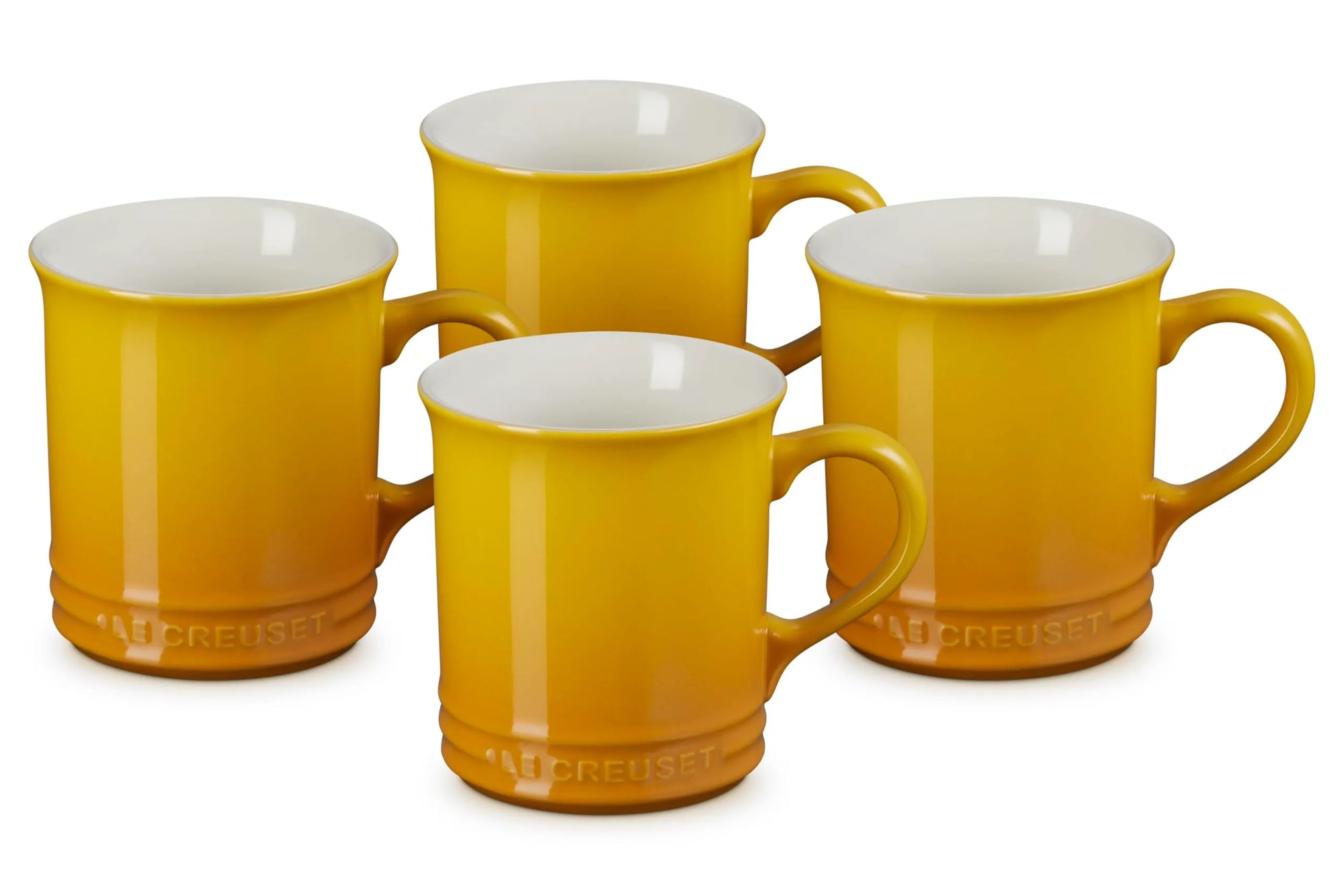 Four Le Creuset ceramic mugs in a warm mustard yellow color with a glossy finish and white interiors, arranged on a clean white background.