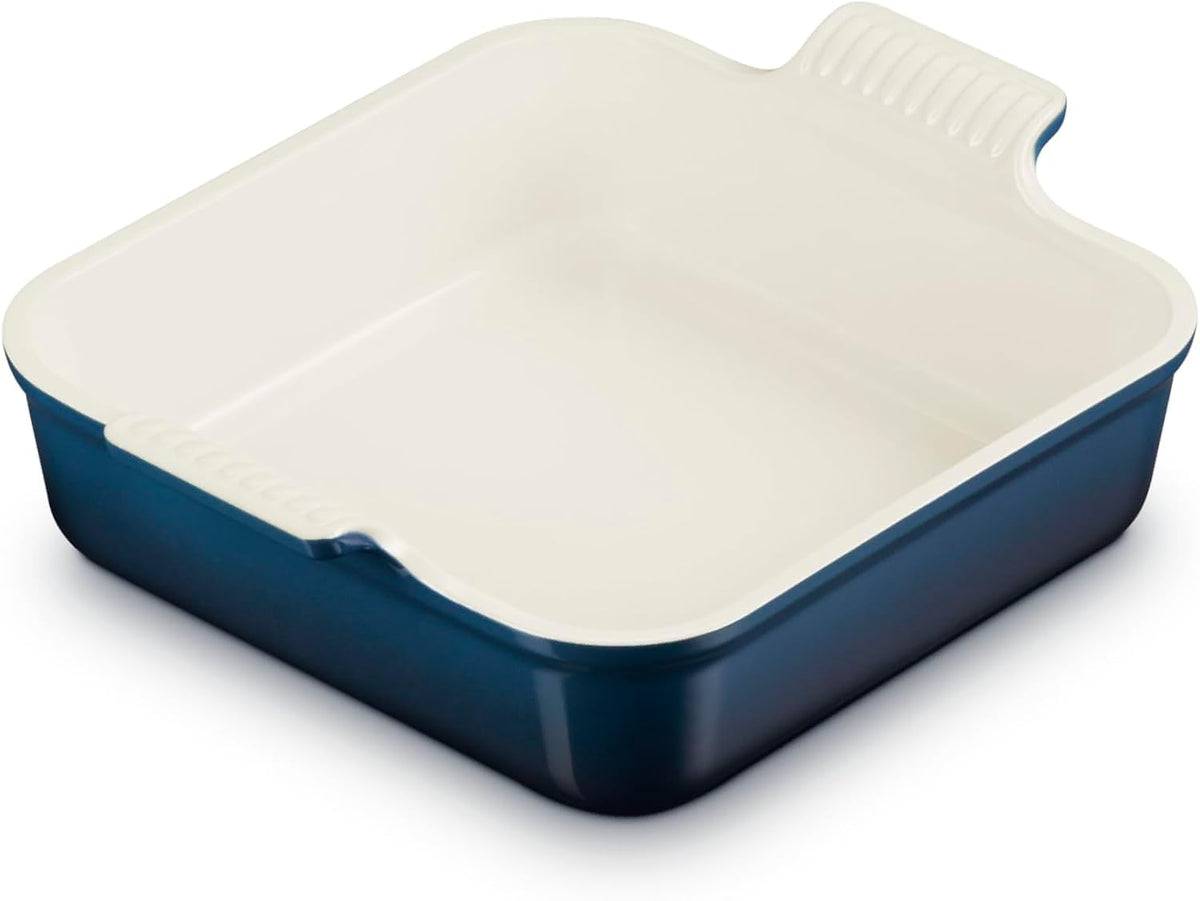 A square ceramic baking dish with a navy blue exterior and a cream-colored interior, featuring a handle on the top right with a ribbed texture.