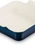 A square ceramic baking dish with a navy blue exterior and a cream-colored interior, featuring a handle on the top right with a ribbed texture.