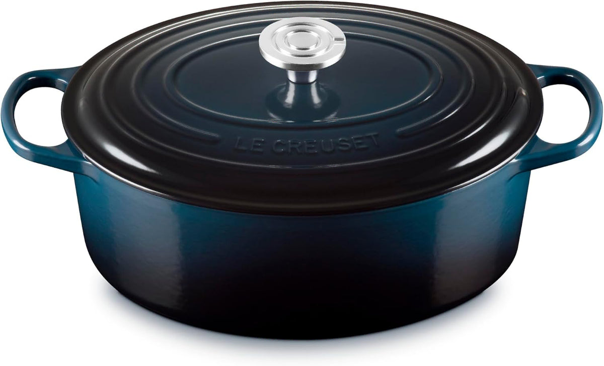 A Le Creuset round enameled cast iron Dutch oven with lid, in a dark blue color, featuring side handles and a silver knob, set against a plain white background.