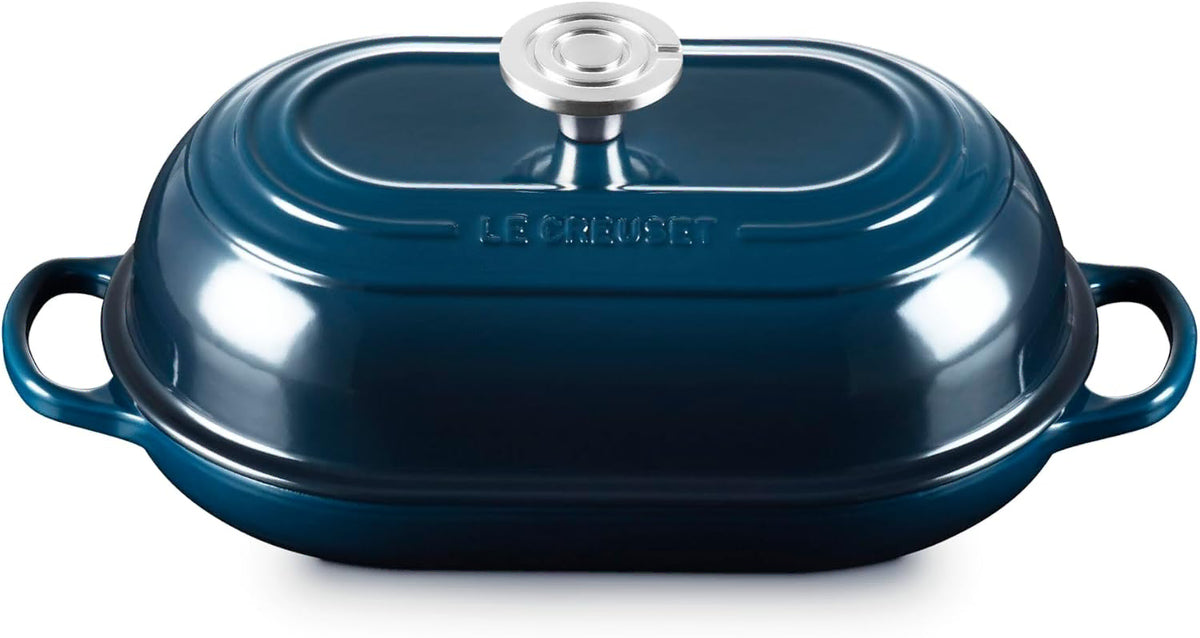 A Le Creuset oval lidded casserole dish in dark teal blue with a silver knob and side handles, shown on a white background.