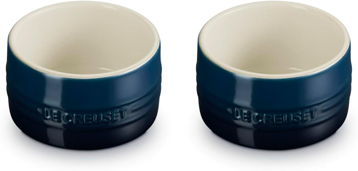 Two Le Creuset ramekins shown side by side on a white background, featuring a dark navy blue exterior with a cream-colored interior.