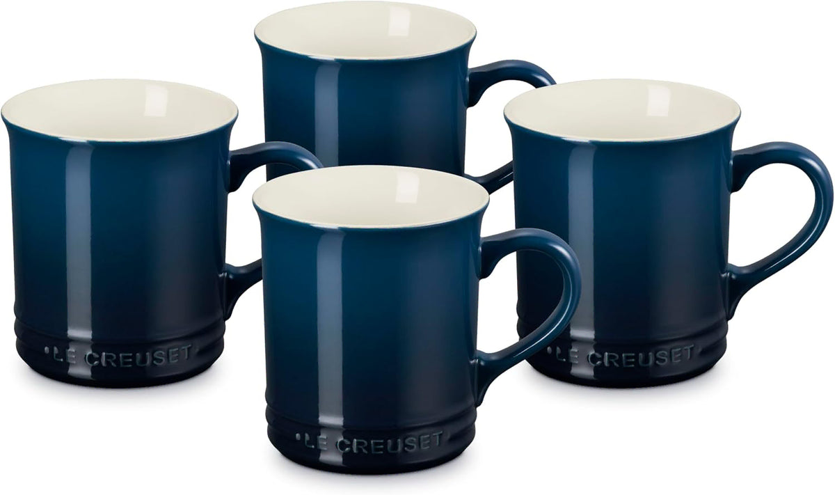 Four Le Creuset dark blue ceramic mugs with cream-colored interiors, arranged together on a white background.