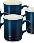 Four Le Creuset dark blue ceramic mugs with cream-colored interiors, arranged together on a white background.