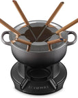 A Le Creuset fondue pot set on a matching black base, with six wooden-handled utensils arranged around the pot on a white background.