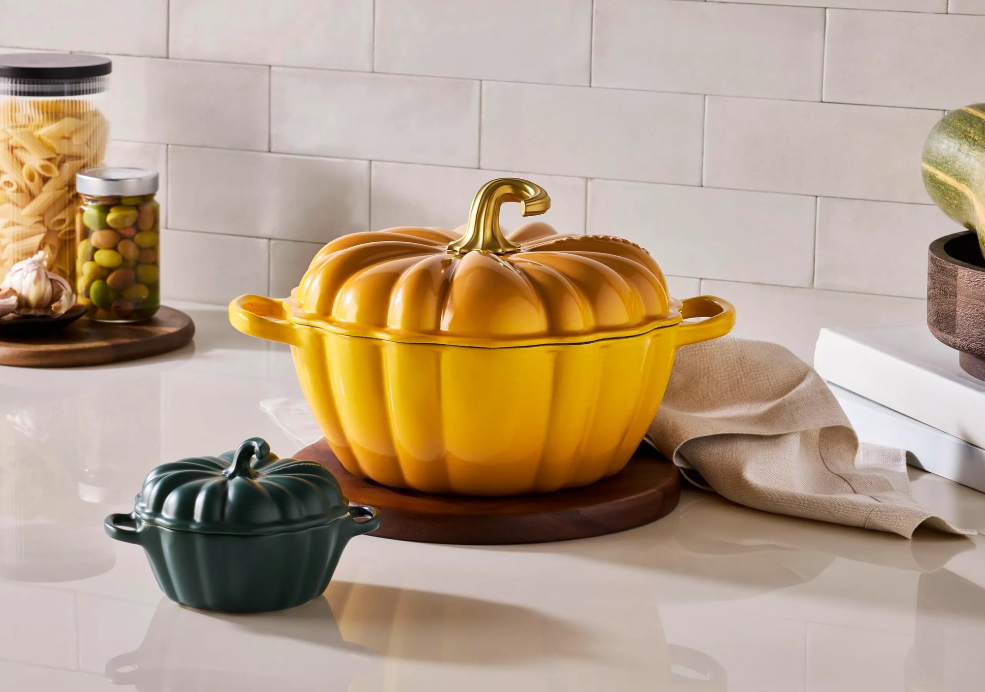 A yellow pumpkin-shaped lidded casserole dish with a gold-toned stem handle sits on a wooden trivet on a kitchen counter, alongside a small dark green pumpkin dish and kitchen props.