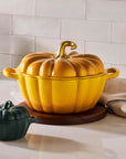 A yellow pumpkin-shaped lidded casserole dish with a gold-toned stem handle sits on a wooden trivet on a kitchen counter, alongside a small dark green pumpkin dish and kitchen props.