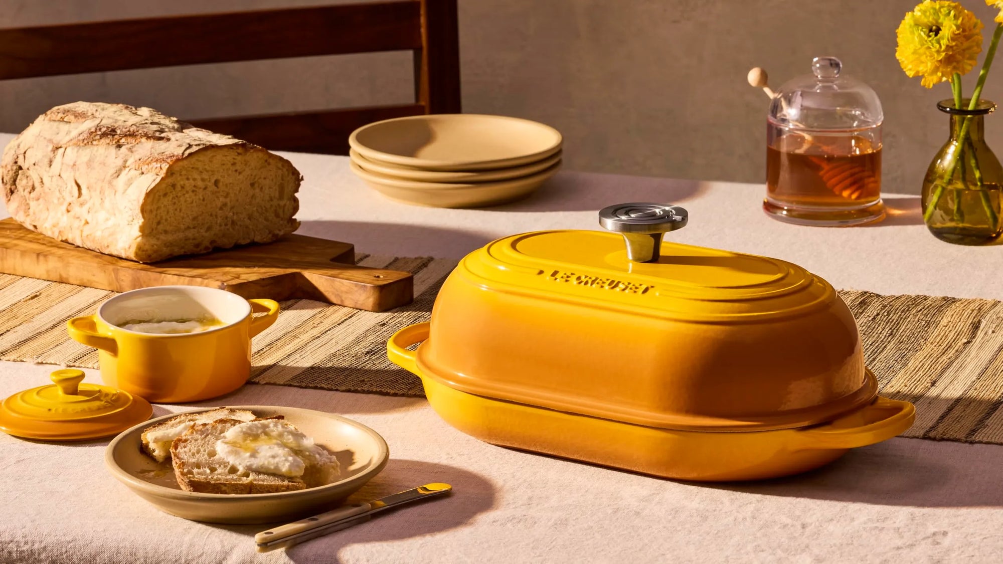A yellow enameled cast iron casserole dish with a lid sits on a dining table styled with a loaf of bread, a small yellow bowl, plates, and a glass jar of honey in warm natural light.