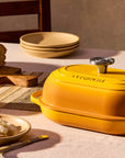A yellow enameled cast iron casserole dish with a lid sits on a dining table styled with a loaf of bread, a small yellow bowl, plates, and a glass jar of honey in warm natural light.