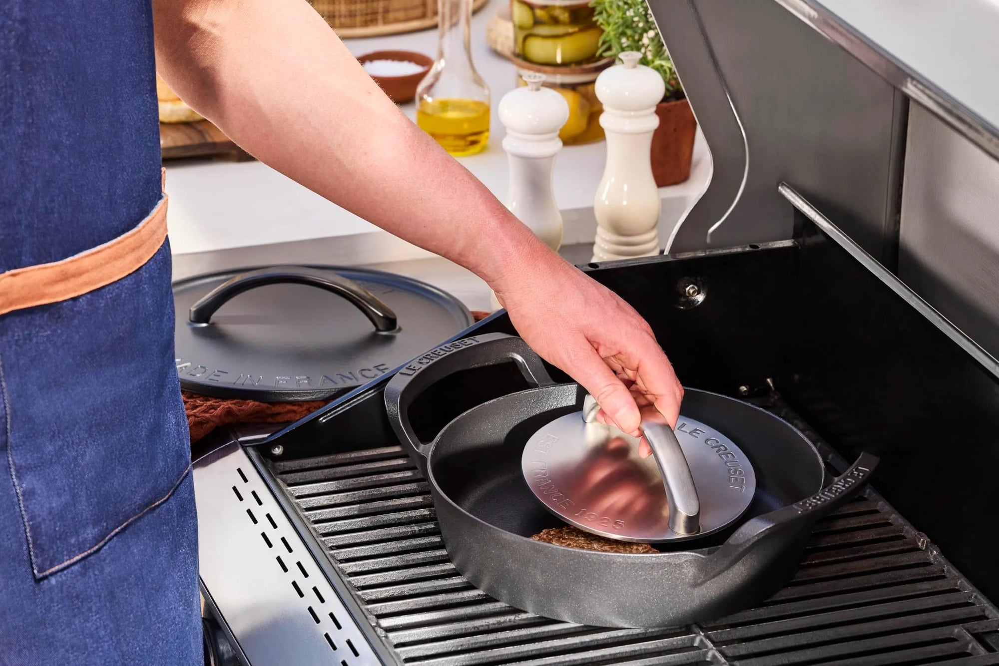 A person in a blue apron uses a grill press to press a meat patty on a cast iron skillet from a grill, with another skillet and kitchen items visible in the background.