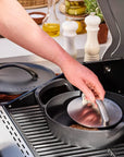 A person in a blue apron uses a grill press to press a meat patty on a cast iron skillet from a grill, with another skillet and kitchen items visible in the background.