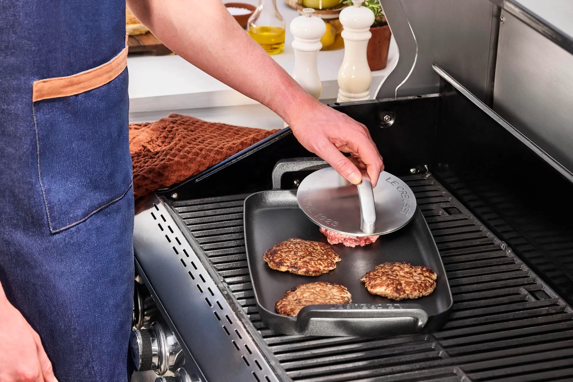 A person wearing a blue apron uses a round metal grill press with handle to press a cooked burger patty from a black square grill pan on a gas grill.