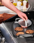 A person wearing a blue apron uses a round metal grill press with handle to press a cooked burger patty from a black square grill pan on a gas grill.