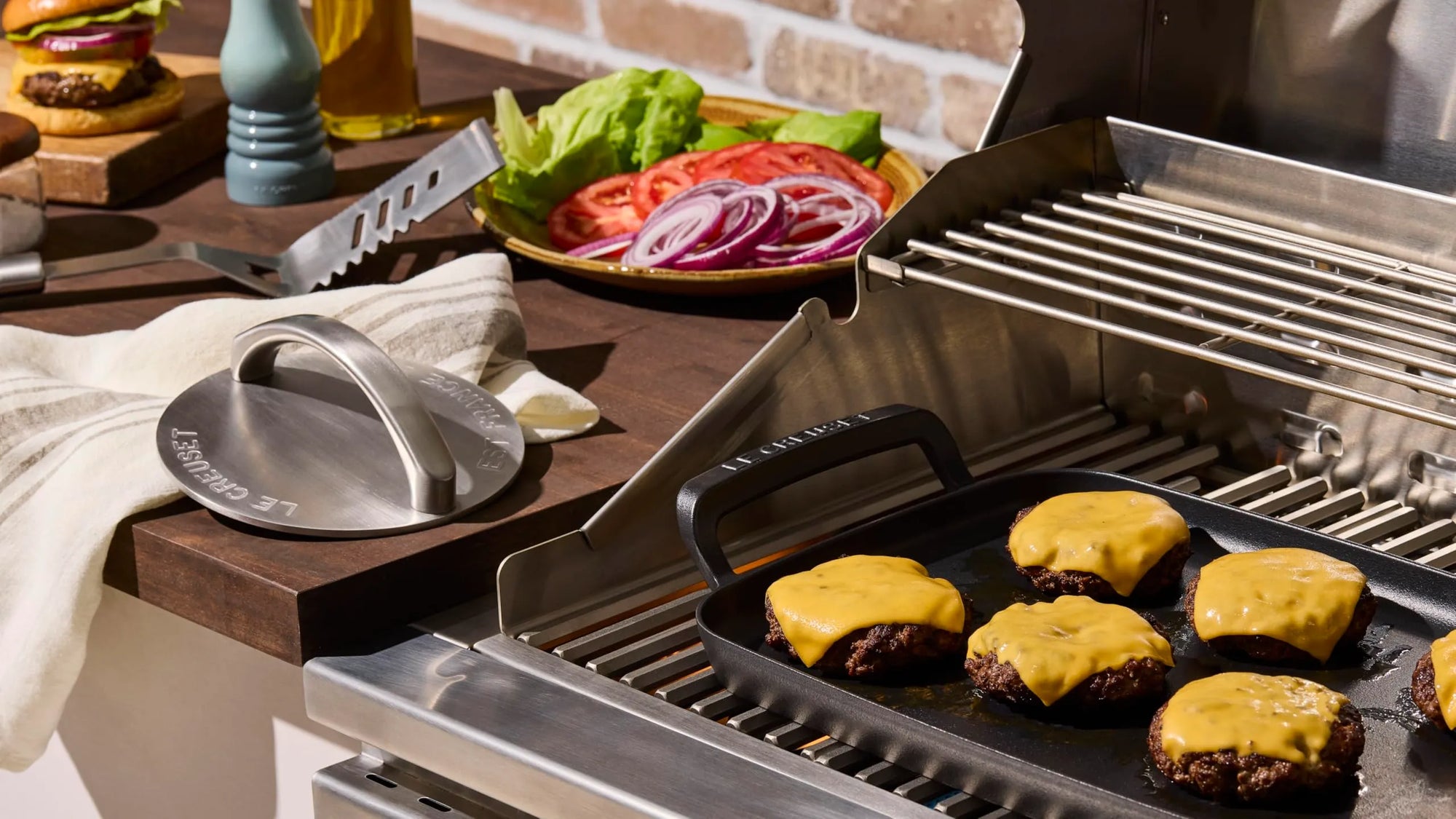 A stainless steel outdoor grill with cheese-covered beef patties cooking on the grates, alongside a grill press, a towel, and a plate of sliced vegetables in a kitchen setting.
