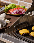 A stainless steel outdoor grill with cheese-covered beef patties cooking on the grates, alongside a grill press, a towel, and a plate of sliced vegetables in a kitchen setting.