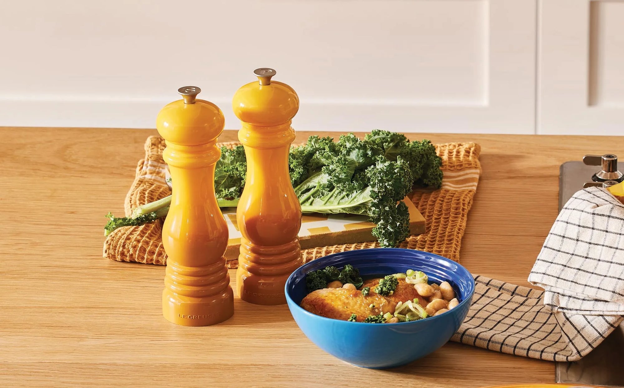 Two yellow ceramic pepper mills standing on a wooden countertop next to a blue bowl of food, with kale on a woven mat in the background.