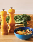 Two yellow ceramic pepper mills standing on a wooden countertop next to a blue bowl of food, with kale on a woven mat in the background.