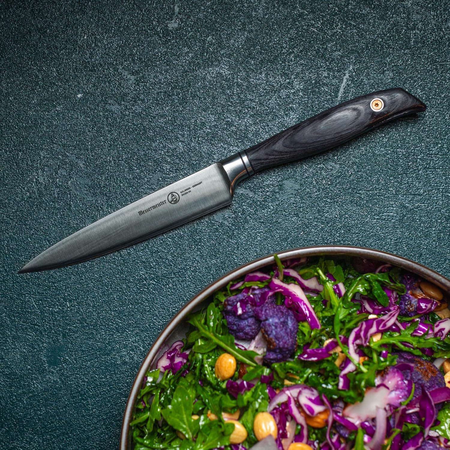 Utility knife with black handle on a dark surface next to a salad bowl with a salad.