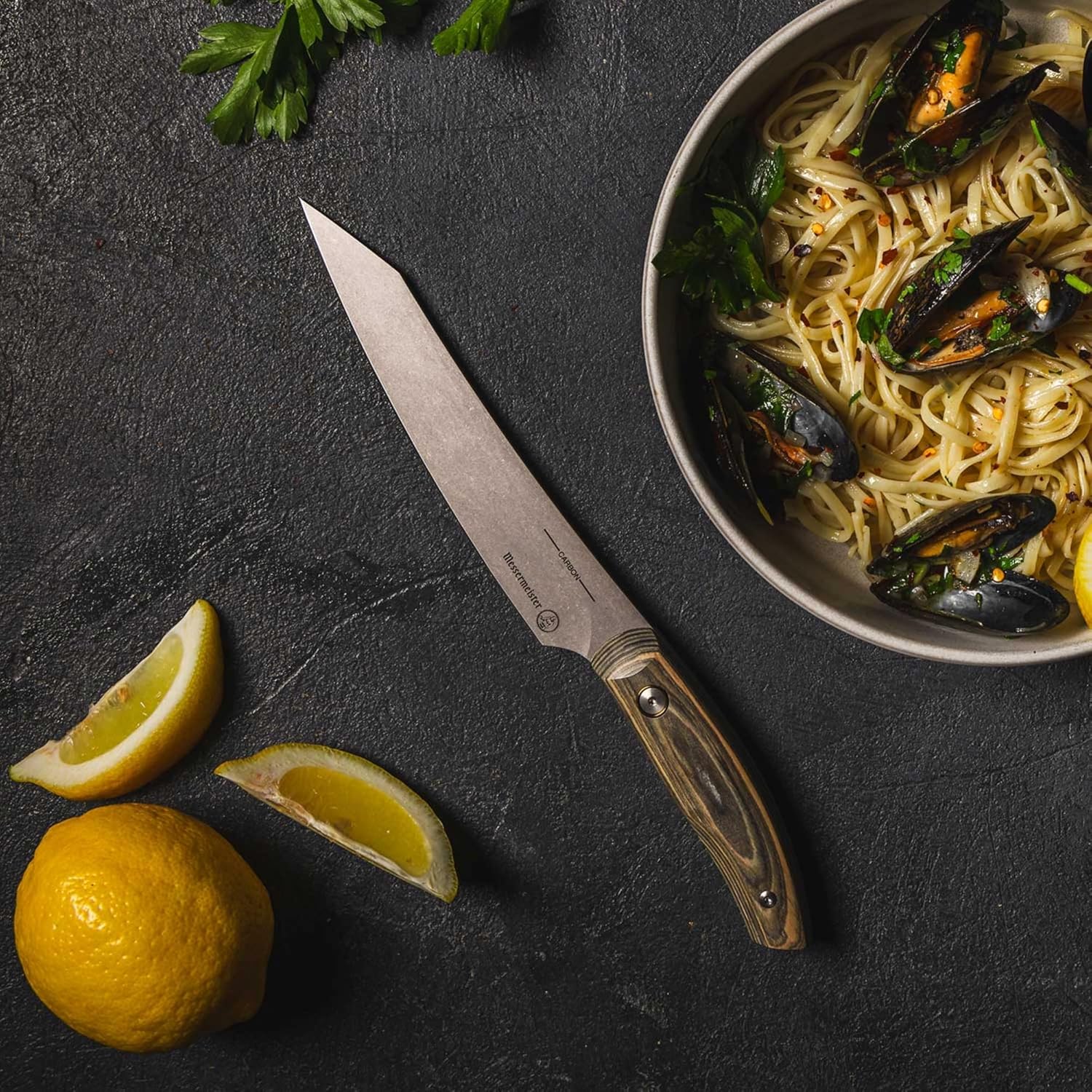 Knife on a dark surface with a bowl of pasta and mussels, lemon wedges, and parsley.