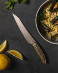 Knife on a dark surface with a bowl of pasta and mussels, lemon wedges, and parsley.