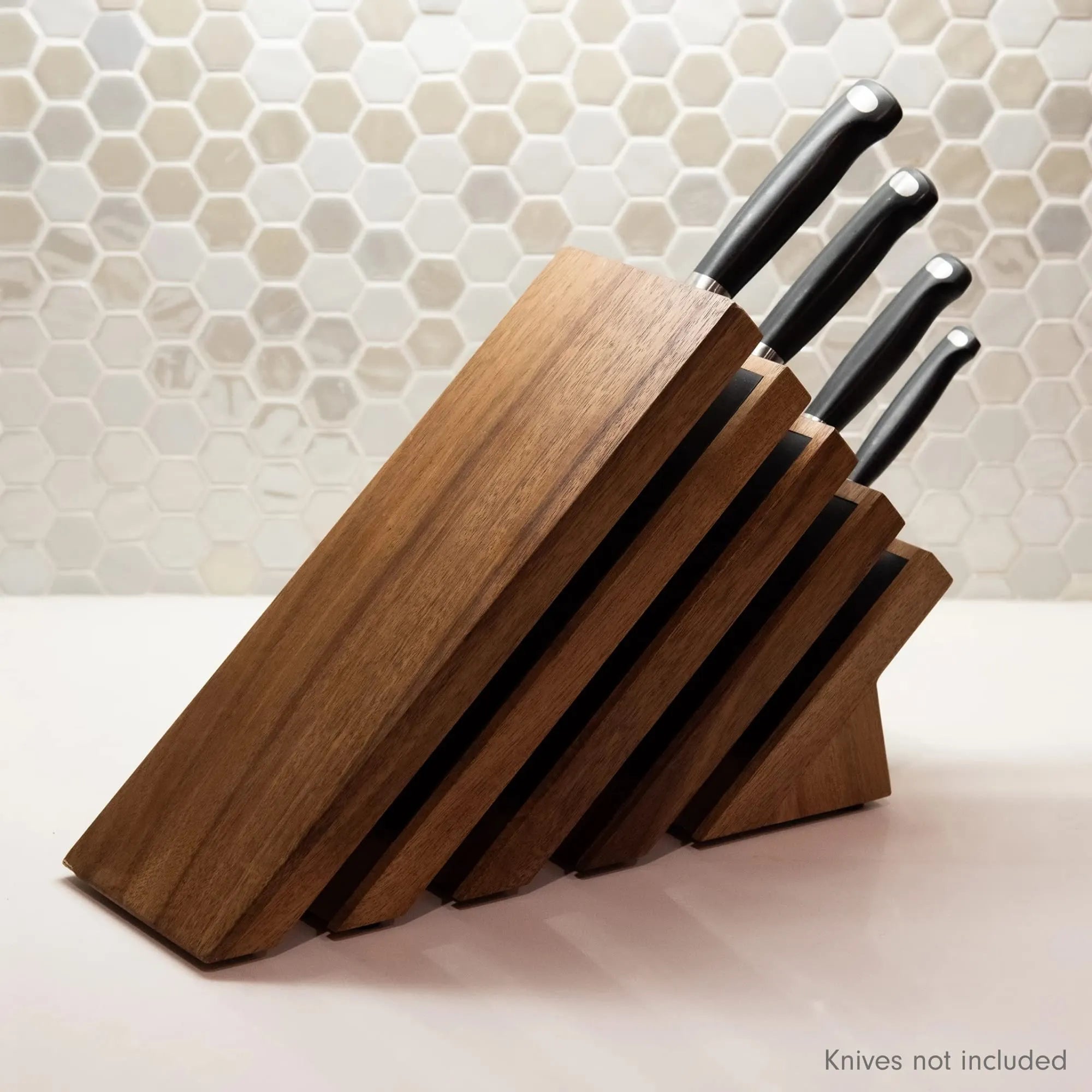 A wooden knife block with angled slots holding four knives with black handles, set on a light countertop in front of a beige hexagonal tile backsplash. Text in the bottom right corner reads 'Knives not included'.