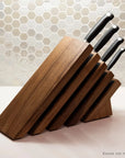 A wooden knife block with angled slots holding four knives with black handles, set on a light countertop in front of a beige hexagonal tile backsplash. Text in the bottom right corner reads 'Knives not included'.