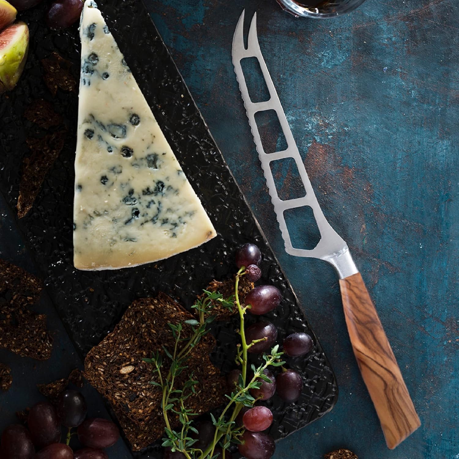 Triangle of blue cheese with a cheese knife on a dark surface with grapes and bread.