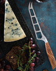 Triangle of blue cheese with a cheese knife on a dark surface with grapes and bread.