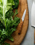 A chef knife and a pairing knife with wooden handles on a wooden cutting board next to fresh green leafy vegetables.