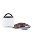 White coffee canister with black lid and handle, surrounded by coffee beans on a white background.