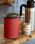 Red container, silver thermos, and spice jars on a wooden surface with a blurred background.