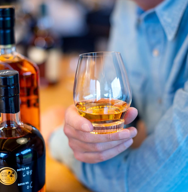 A person in a light blue shirt holds a clear tulip-style whiskey glass with amber liquor, with two dark whiskey bottles blurred in the foreground and background on a warm bar counter.