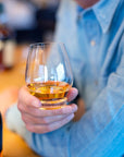 Person holding a glass of bourbon with bottles in the background