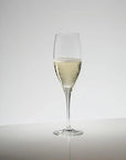 A clear champagne flute glass partially filled with pale golden bubbly beverage, centered on a light gray studio background with a reflective surface below.