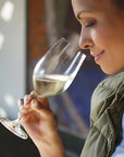 A woman in profile holding a wine glass with white wine, wearing an olive green jacket over a white top, in a softly lit indoor setting.